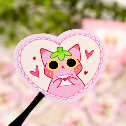 Heart-shaped pink object with a cartoon cat design held by a hand against a blurred floral background